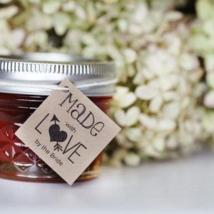 Puede incluir: Un peque&ntilde;o tarro de vidrio lleno de mermelada roja, con una etiqueta de papel marr&oacute;n que dice "Made with LOVE by the Bride".