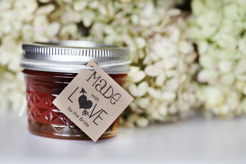 Puede incluir: Un peque&ntilde;o tarro de vidrio lleno de mermelada roja, con una etiqueta de papel marr&oacute;n que dice "Made with LOVE by the Bride".