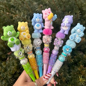 May include: A collection of colorful pens, each topped with a different Care Bear figurine. The pens feature a variety of colors, including green, yellow, blue, pink, and purple, with decorative embellishments and sparkling accents.