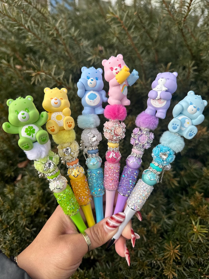 May include: A collection of colorful pens, each topped with a different Care Bear figurine. The pens feature a variety of colors, including green, yellow, blue, pink, and purple, with decorative embellishments and sparkling accents.