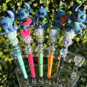 May include: A collection of five pens featuring blue Stitch character toppers. Each pen has a unique design with colorful beads, pom-poms, and jewel accents. The pen barrels are in various colors, including teal, pink, green, orange, and black.