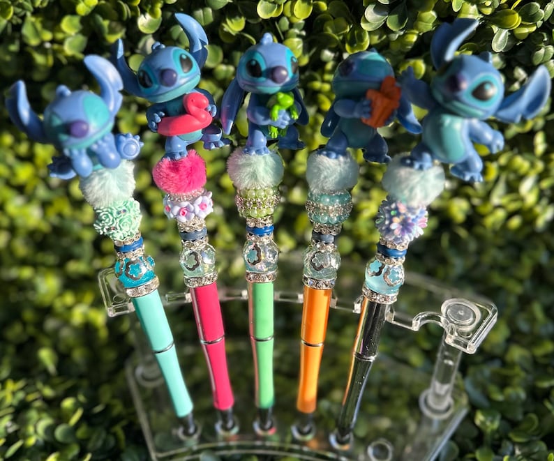 May include: A collection of five pens featuring blue Stitch character toppers. Each pen has a unique design with colorful beads, pom-poms, and jewel accents. The pen barrels are in various colors, including teal, pink, green, orange, and black.