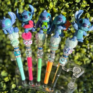 May include: Five colorful pens featuring blue Stitch figurines. Each pen has a unique design with decorative beads, faux fur, and a different colored pen barrel: teal, pink, green, orange, and black. The pens are displayed in a clear plastic holder.