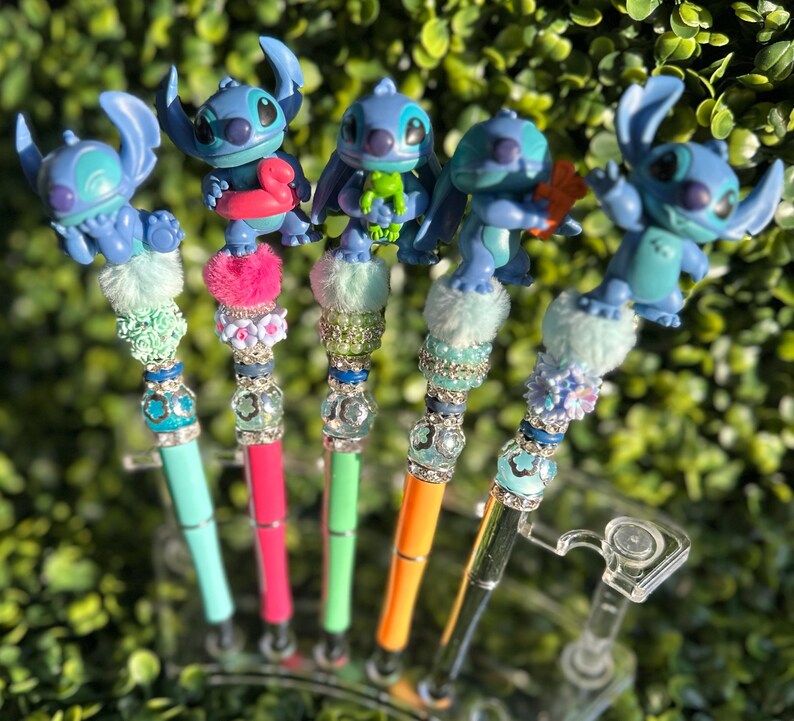 May include: Five colorful pens featuring blue Stitch figurines. Each pen has a unique design with decorative beads, faux fur, and a different colored pen barrel: teal, pink, green, orange, and black. The pens are displayed in a clear plastic holder.