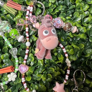 K&ouml;nnte beinhalten: Ein rosa Esel-Schl&uuml;sselanh&auml;nger mit einem Perlenband mit rosa, wei&szlig;en und braunen Perlen, einem rosa Stern und wei&szlig;en Blumenanh&auml;ngern. Der Schl&uuml;sselanh&auml;nger hat einen silbernen Verschluss und einen gr&uuml;nen Hintergrund.