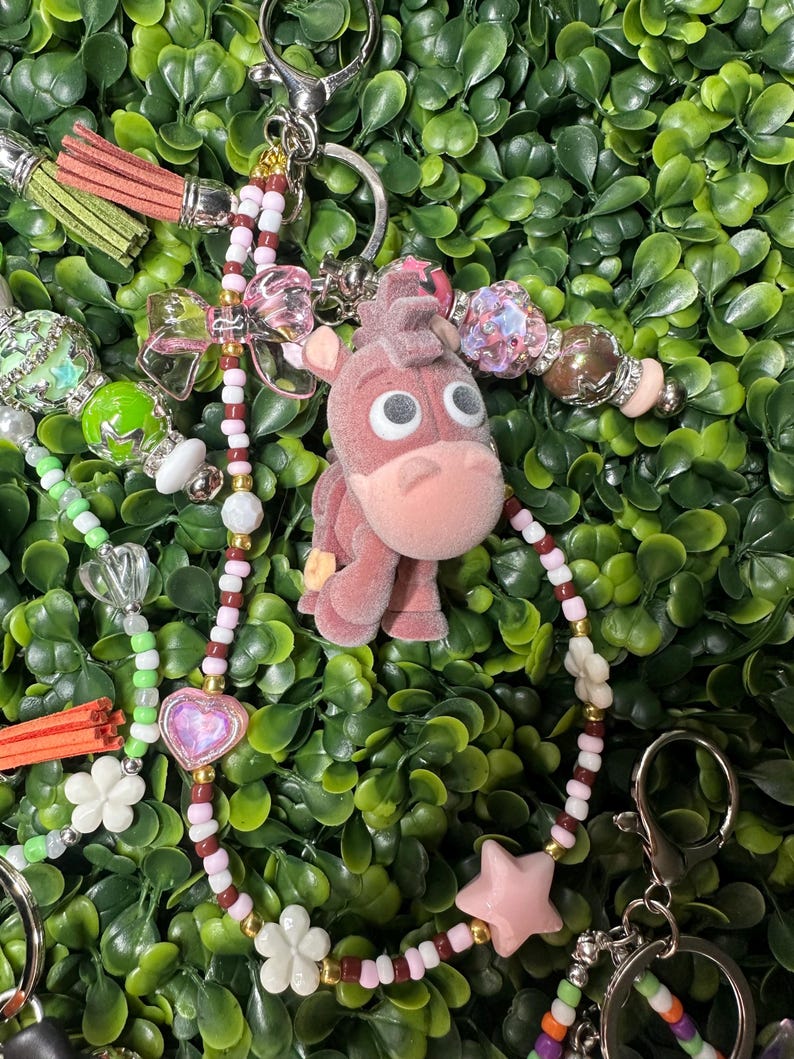 K&ouml;nnte beinhalten: Ein rosa Esel-Schl&uuml;sselanh&auml;nger mit einem Perlenband mit rosa, wei&szlig;en und braunen Perlen, einem rosa Stern und wei&szlig;en Blumenanh&auml;ngern. Der Schl&uuml;sselanh&auml;nger hat einen silbernen Verschluss und einen gr&uuml;nen Hintergrund.