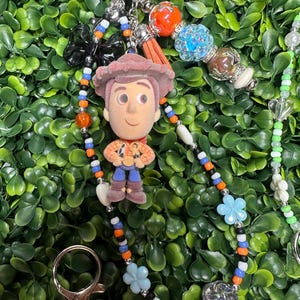 K&ouml;nnte beinhalten: Ein Schl&uuml;sselanh&auml;nger mit einer kleinen Woody-Figur aus Toy Story, mit einem Perlenband in Orange, Blau und Schwarz. Der Schl&uuml;sselanh&auml;nger enth&auml;lt einen silbernen Verschluss, einen Quastenanh&auml;nger und verschiedene dekorative Perlen und Charms.
