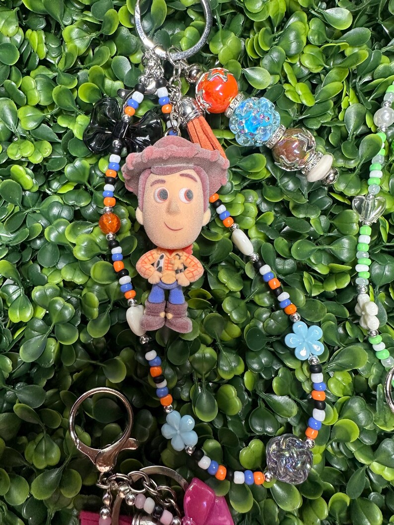K&ouml;nnte beinhalten: Ein Schl&uuml;sselanh&auml;nger mit einer kleinen Woody-Figur aus Toy Story, mit einem Perlenband in Orange, Blau und Schwarz. Der Schl&uuml;sselanh&auml;nger enth&auml;lt einen silbernen Verschluss, einen Quastenanh&auml;nger und verschiedene dekorative Perlen und Charms.