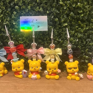 May include: A collection of decorative clips featuring yellow bear figurines with red shirts and various accessories. Each clip has a decorative top with beads and bows in different colors. A card with the text "Thank you" is visible.