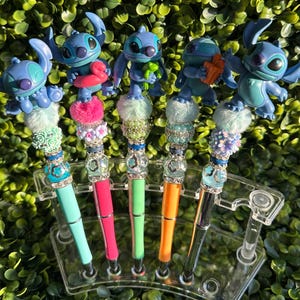 May include: A collection of five decorative pens featuring blue cartoon character figures. Each pen has a different colored barrel, including teal, pink, green, orange, and black. The pens are embellished with decorative beads and fluffy accents.