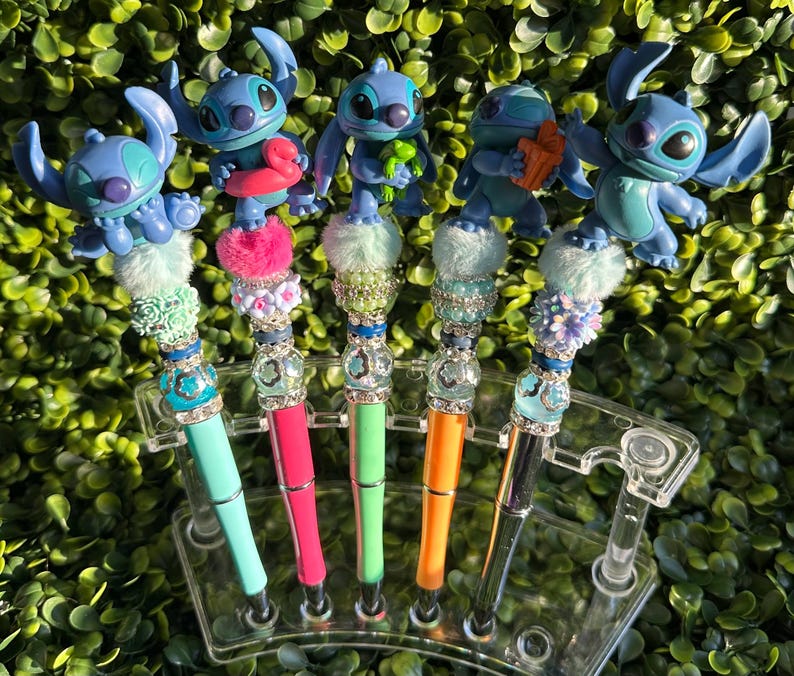 May include: A collection of five decorative pens featuring blue cartoon character figures. Each pen has a different colored barrel, including teal, pink, green, orange, and black. The pens are embellished with decorative beads and fluffy accents.