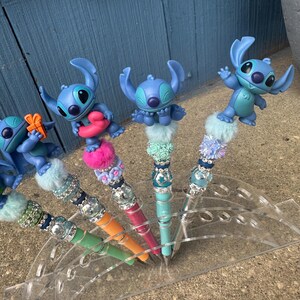 May include: A collection of decorative pens featuring blue cartoon character figures. The pens have colorful barrels in green, orange, pink, and teal, adorned with sparkling embellishments and fluffy accents. The pens are displayed in a clear acrylic holder.