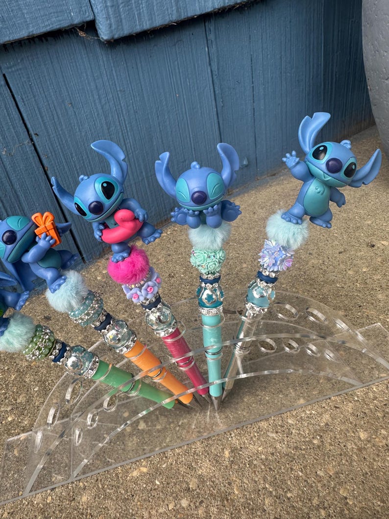 May include: A collection of decorative pens featuring blue cartoon character figures. The pens have colorful barrels in green, orange, pink, and teal, adorned with sparkling embellishments and fluffy accents. The pens are displayed in a clear acrylic holder.