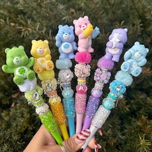 May include: A collection of colorful pens, each topped with a Care Bear figurine. The pens feature various colors, including green, yellow, blue, pink, and purple, with decorative embellishments and sparkling accents. The pens are held in a hand.