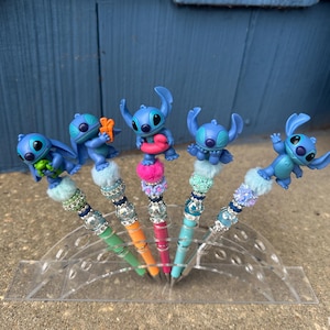 May include: Five pens featuring blue Stitch figures on top. The pens have colorful barrels in green, orange, pink, and blue. Each pen has a decorative top with beads and faux fur. The pens are displayed in a clear acrylic holder.