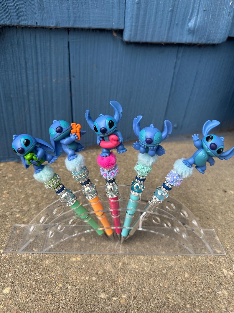 May include: Five pens featuring blue Stitch figures on top. The pens have colorful barrels in green, orange, pink, and blue. Each pen has a decorative top with beads and faux fur. The pens are displayed in a clear acrylic holder.