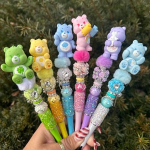 May include: A collection of colorful pens, each adorned with a different Care Bear figurine. The pens feature various colors, including green, yellow, blue, pink, and purple, with decorative embellishments and sparkling accents. The pens are held together in a hand.