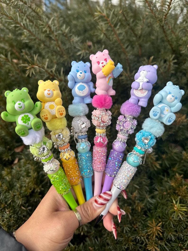 May include: A collection of colorful pens, each adorned with a different Care Bear figurine. The pens feature various colors, including green, yellow, blue, pink, and purple, with decorative embellishments and sparkling accents. The pens are held together in a hand.