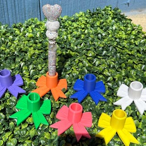 May include: A bedazzled wand with a heart-shaped top and a silver, crystal-studded shaft. Six colorful bow-shaped candle holders in purple, orange, blue, green, pink, white, and yellow are arranged around the wand. The items are set on a bed of green foliage.