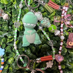 K&ouml;nnte beinhalten: Eine Sammlung von Schl&uuml;sselanh&auml;ngern mit verschiedenen Anh&auml;ngern. Ein Schl&uuml;sselanh&auml;nger hat einen gr&uuml;nen Dinosaurier-Anh&auml;nger. Andere Schl&uuml;sselanh&auml;nger haben Herz-, Blumen- und Schleifenanh&auml;nger. Die Schl&uuml;sselanh&auml;nger haben silberne Verschl&uuml;sse und bunte Perlen, einschlie&szlig;lich Perlen und Quasten.