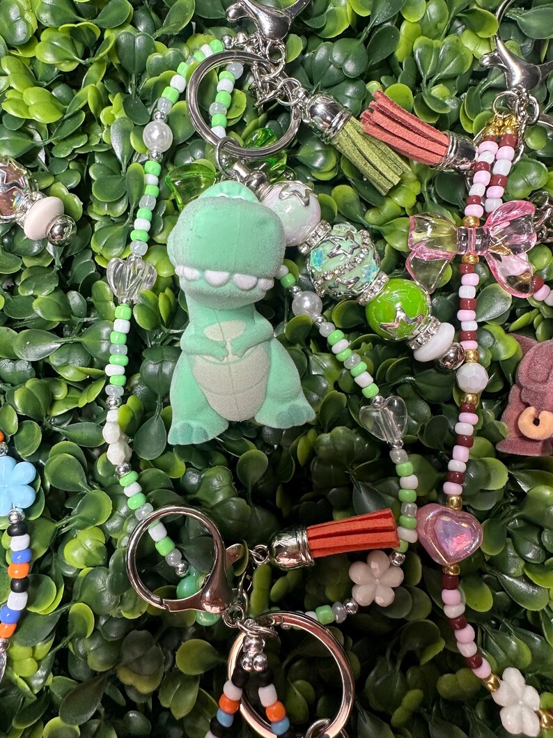 K&ouml;nnte beinhalten: Eine Sammlung von Schl&uuml;sselanh&auml;ngern mit verschiedenen Anh&auml;ngern. Ein Schl&uuml;sselanh&auml;nger hat einen gr&uuml;nen Dinosaurier-Anh&auml;nger. Andere Schl&uuml;sselanh&auml;nger haben Herz-, Blumen- und Schleifenanh&auml;nger. Die Schl&uuml;sselanh&auml;nger haben silberne Verschl&uuml;sse und bunte Perlen, einschlie&szlig;lich Perlen und Quasten.
