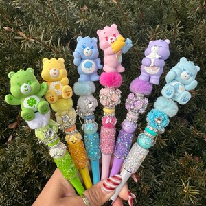 May include: A collection of colorful pens featuring Care Bear figurines. Each pen has a different Care Bear character on top, with decorative embellishments and sparkling accents. The pens are held in a hand against a backdrop of green foliage.