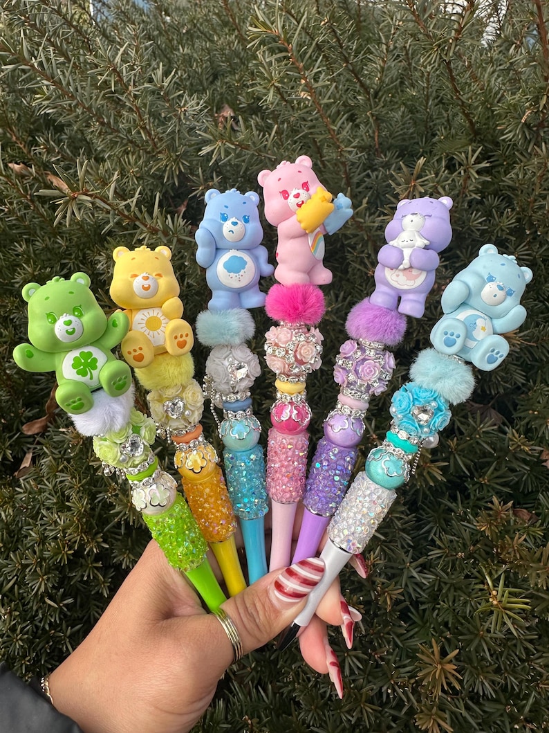 May include: A collection of colorful pens featuring Care Bear figurines. Each pen has a different Care Bear character on top, with decorative embellishments and sparkling accents. The pens are held in a hand against a backdrop of green foliage.