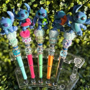 May include: A collection of five decorative pens featuring blue alien figures. Each pen has a different colored barrel, including teal, pink, green, orange, and black. The pens are adorned with decorative beads and fluffy accents, and are displayed in a clear plastic holder.