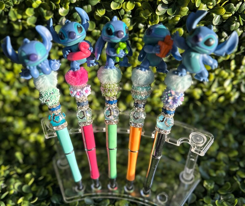 May include: A collection of five decorative pens featuring blue alien figures. Each pen has a different colored barrel, including teal, pink, green, orange, and black. The pens are adorned with decorative beads and fluffy accents, and are displayed in a clear plastic holder.