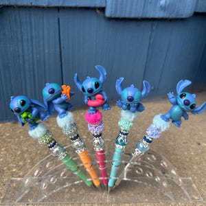 May include: Five pens featuring blue character figures. Each pen has a different colored barrel: green, orange, pink, and blue. The pens are decorated with faux fur and rhinestones. The pens are displayed in a clear plastic holder.