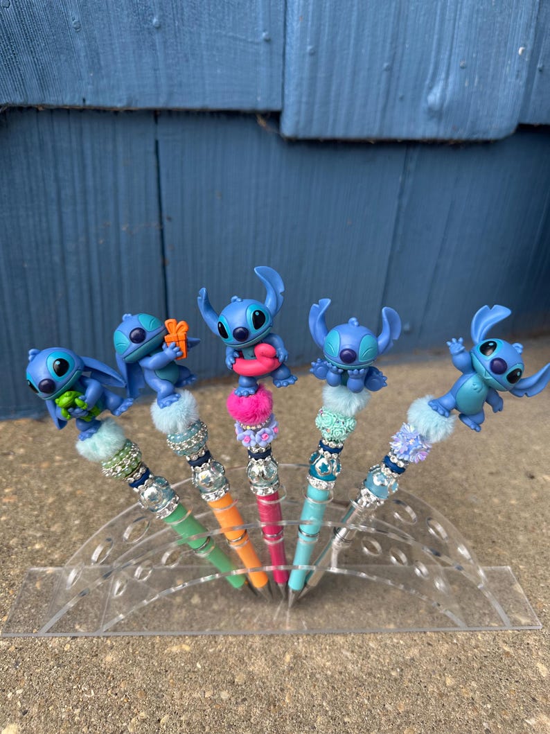 May include: Five pens featuring blue character figures. Each pen has a different colored barrel: green, orange, pink, and blue. The pens are decorated with faux fur and rhinestones. The pens are displayed in a clear plastic holder.
