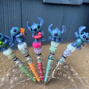 May include: Five decorative pens featuring blue cartoon character figures. Each pen has a different design, with colorful beads, faux fur, and a unique Stitch figurine on top. The pens are displayed in a clear plastic holder.