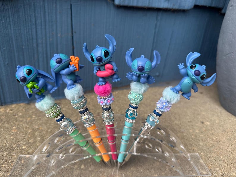 May include: Five decorative pens featuring blue cartoon character figures. Each pen has a different design, with colorful beads, faux fur, and a unique Stitch figurine on top. The pens are displayed in a clear plastic holder.