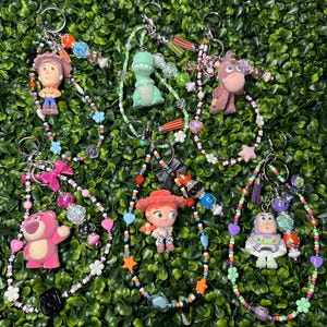 K&ouml;nnte beinhalten: Eine Sammlung von Schl&uuml;sselanh&auml;ngern mit Perlen, die verschiedene Toy Story-Charaktere zeigen. Jeder Schl&uuml;sselanh&auml;nger hat einen anderen Charakter, darunter Woody, Rex, Bullseye, Lotso, Jessie und Buzz Lightyear, mit bunten Perlen und Charms.