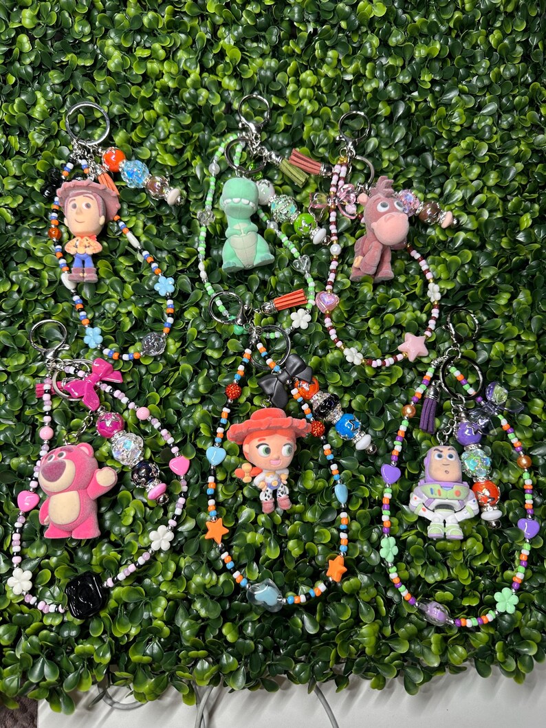 K&ouml;nnte beinhalten: Eine Sammlung von Schl&uuml;sselanh&auml;ngern mit Perlen, die verschiedene Toy Story-Charaktere zeigen. Jeder Schl&uuml;sselanh&auml;nger hat einen anderen Charakter, darunter Woody, Rex, Bullseye, Lotso, Jessie und Buzz Lightyear, mit bunten Perlen und Charms.
