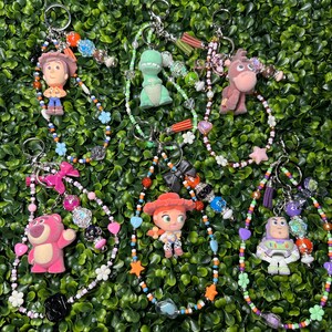 K&ouml;nnte beinhalten: Schl&uuml;sselanh&auml;nger mit verschiedenen Toy Story-Charakteren, darunter Woody, Rex und Buzz Lightyear. Jeder Schl&uuml;sselanh&auml;nger hat ein Perlenband mit bunten Perlen, Charms und einem Metallring. Die Schl&uuml;sselanh&auml;nger sind vor einem gr&uuml;nen Hintergrund.
