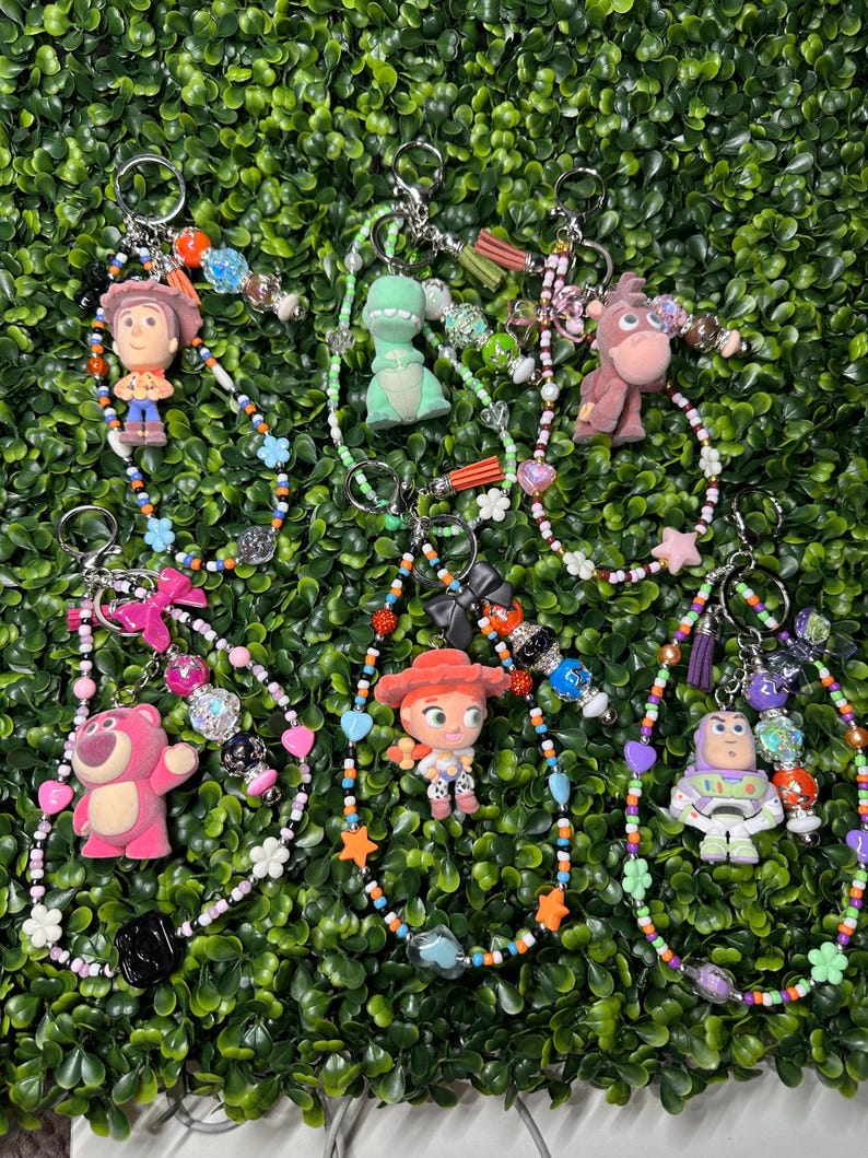 K&ouml;nnte beinhalten: Schl&uuml;sselanh&auml;nger mit verschiedenen Toy Story-Charakteren, darunter Woody, Rex und Buzz Lightyear. Jeder Schl&uuml;sselanh&auml;nger hat ein Perlenband mit bunten Perlen, Charms und einem Metallring. Die Schl&uuml;sselanh&auml;nger sind vor einem gr&uuml;nen Hintergrund.