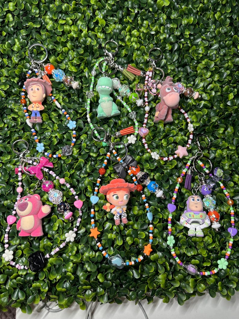 K&ouml;nnte beinhalten: Schl&uuml;sselanh&auml;nger mit Charakteren aus dem Toy Story-Franchise. Jeder Schl&uuml;sselanh&auml;nger hat einen Charakteranh&auml;nger, wie Woody, Rex und Buzz Lightyear, mit bunten Perlenakzenten und einem Metallring. Die Schl&uuml;sselanh&auml;nger werden vor einem gr&uuml;nen Hintergrund pr&auml;sentiert.