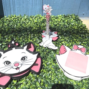May include: White cat-themed desk accessories, including a pen with a cat figurine, a notepad holder, and a decorative cat face. The accessories feature pink accents and black outlines, set against a green backdrop.