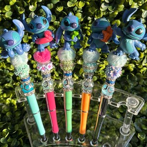 May include: A collection of decorative pens featuring blue cartoon character figurines. Each pen has a unique design with colorful accents, including pom-poms, beads, and rhinestones. The pens are displayed in a clear plastic holder against a backdrop of green foliage.
