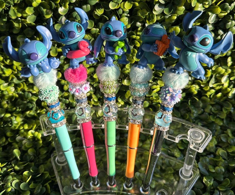 May include: A collection of decorative pens featuring blue cartoon character figurines. Each pen has a unique design with colorful accents, including pom-poms, beads, and rhinestones. The pens are displayed in a clear plastic holder against a backdrop of green foliage.
