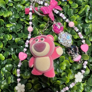 K&ouml;nnte beinhalten: Ein pinkfarbener Schl&uuml;sselanh&auml;nger mit einem Pl&uuml;schb&auml;ren-Charakter, einer pinkfarbenen Schleife und verschiedenen Perlen in Pink, Wei&szlig;, Schwarz und Silber. Der Schl&uuml;sselanh&auml;nger hat einen silbernen Verschluss und einen schwarzen quadratischen Anh&auml;nger, vor gr&uuml;nem Laub.