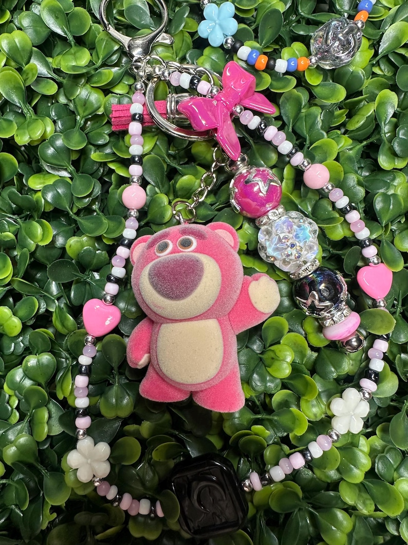 K&ouml;nnte beinhalten: Ein pinkfarbener Schl&uuml;sselanh&auml;nger mit einem Pl&uuml;schb&auml;ren-Charakter, einer pinkfarbenen Schleife und verschiedenen Perlen in Pink, Wei&szlig;, Schwarz und Silber. Der Schl&uuml;sselanh&auml;nger hat einen silbernen Verschluss und einen schwarzen quadratischen Anh&auml;nger, vor gr&uuml;nem Laub.