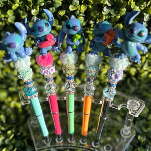 May include: A collection of decorative pens featuring blue alien character toppers. Each pen has a unique design with colorful accents, including pom-poms, beads, and rhinestones. The pens are displayed in a clear plastic holder against a backdrop of green foliage.