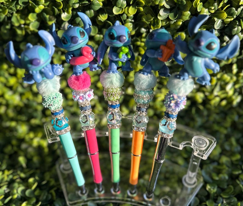 May include: A collection of decorative pens featuring blue alien character toppers. Each pen has a unique design with colorful accents, including pom-poms, beads, and rhinestones. The pens are displayed in a clear plastic holder against a backdrop of green foliage.