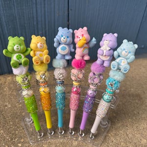 May include: A set of seven colorful pens, each topped with a Care Bear figurine. The pens feature glittery barrels in various colors, including green, yellow, blue, pink, purple, and white. The Care Bears are in different poses and colors.