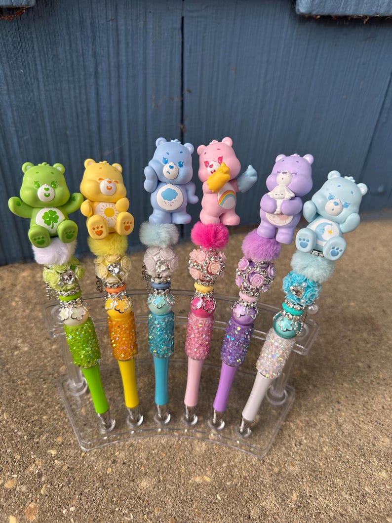 May include: A set of seven colorful pens, each topped with a Care Bear figurine. The pens feature glittery barrels in various colors, including green, yellow, blue, pink, purple, and white. The Care Bears are in different poses and colors.