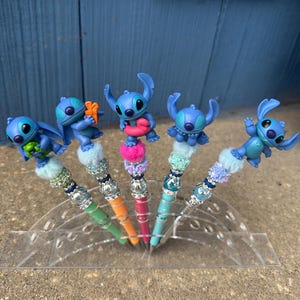 May include: Five pens with blue Stitch character toppers. The pens have colorful barrels in green, orange, pink, and blue. Each pen has a decorative beaded section and a fluffy pom-pom base. The pens are displayed in a clear plastic holder.