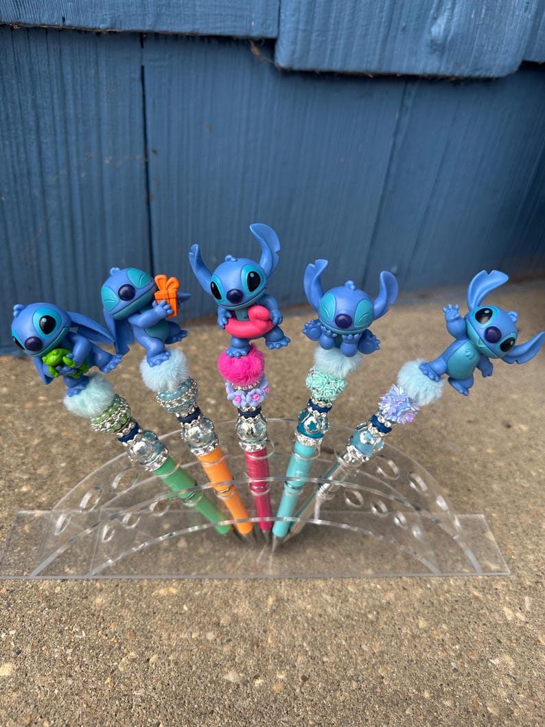 May include: Five pens with blue Stitch character toppers. The pens have colorful barrels in green, orange, pink, and blue. Each pen has a decorative beaded section and a fluffy pom-pom base. The pens are displayed in a clear plastic holder.