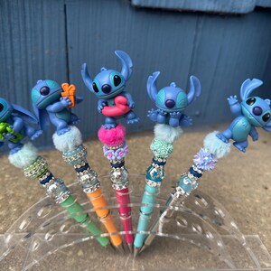 May include: Five decorative pens featuring blue alien character figures. Each pen has a different colored barrel, including green, orange, and pink. The pens are embellished with decorative beads and fluffy accents, and are displayed in a clear plastic holder.